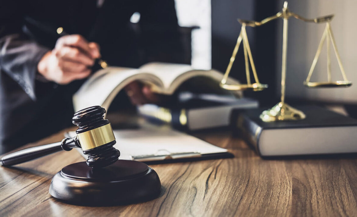 Man writing on a book with books, justice scale and court gravel on a table.