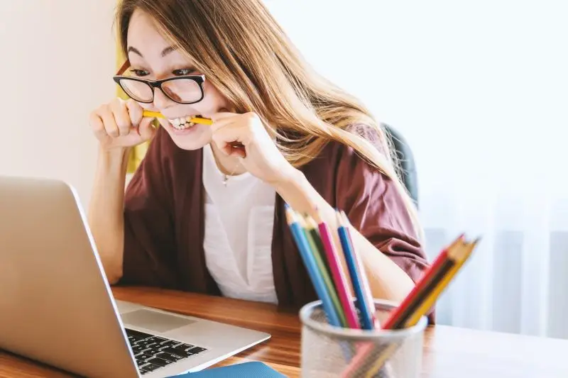 Woman with glasses frustrated biting pencil in her mouth while looking at laptop.