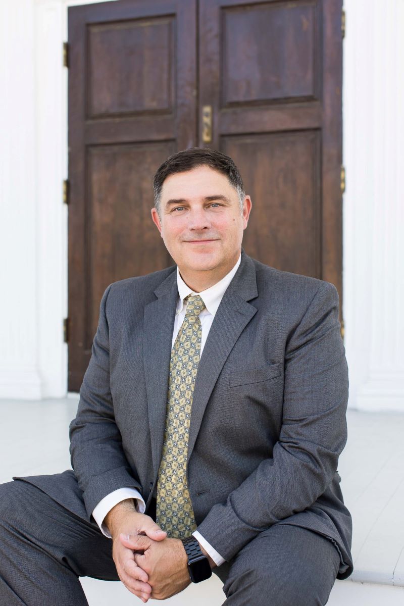 Marc LeBeau portrait in grey suit smiling to the camera while sitting on the floor in front of the door.