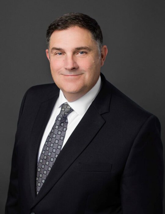 Dr. Marc LeBeau posing professionally smiling in front of the camera in a black suit.